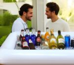 Two white BCWATERFUNPD inflatable serving bars filled with ice, drinks, and snacks at an outdoor party setting.