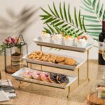 A three-tier Gomakren serving stand with gold frame and white rectangular porcelain platters, displaying various desserts.